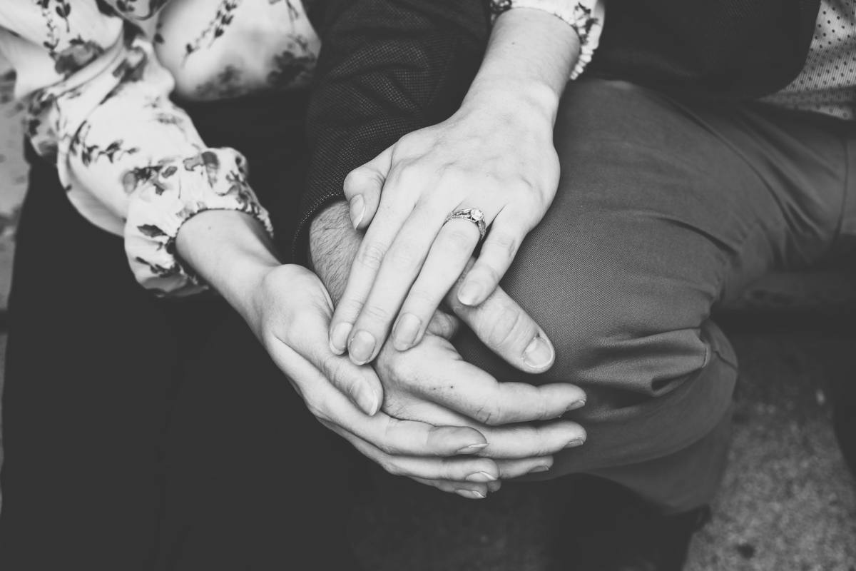 Two people's hands clasped together over a knee, one wearing a wedding ring, in black and white